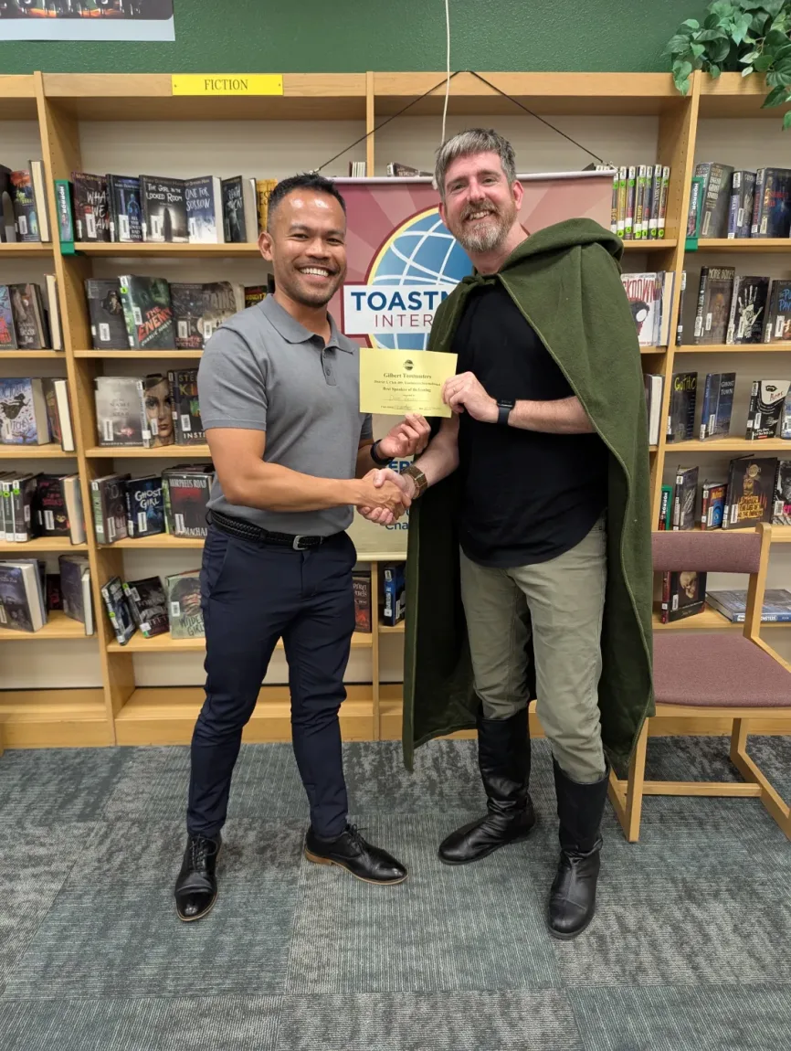 A Gilbert Toastmasters member in a costume cape receiving the Best Speaker certificate with a handshake