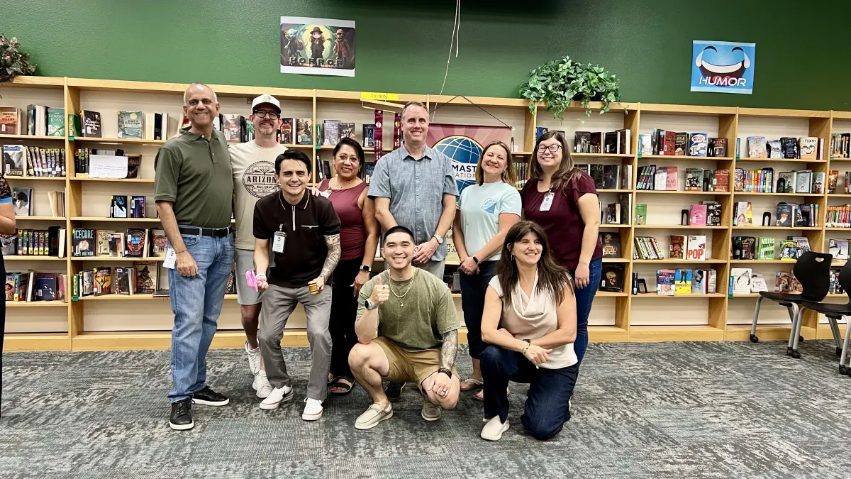 Gilbert Toastmasters officers standing and kneeling together in front of the library bookshelves after the 2026 officer induction ceremony