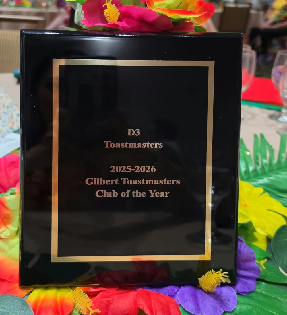 The District 3 Toastmasters Club of the Year plaque for 2025-2026, engraved for Gilbert Toastmasters, standing on a table among silk flowers from the evening's Aloha theme