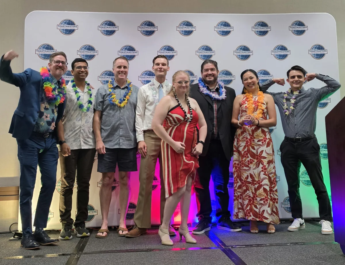 Eight Gilbert Toastmasters members on stage at the Toasties, District 3's annual awards ceremony, wearing leis in front of a Toastmasters International banner