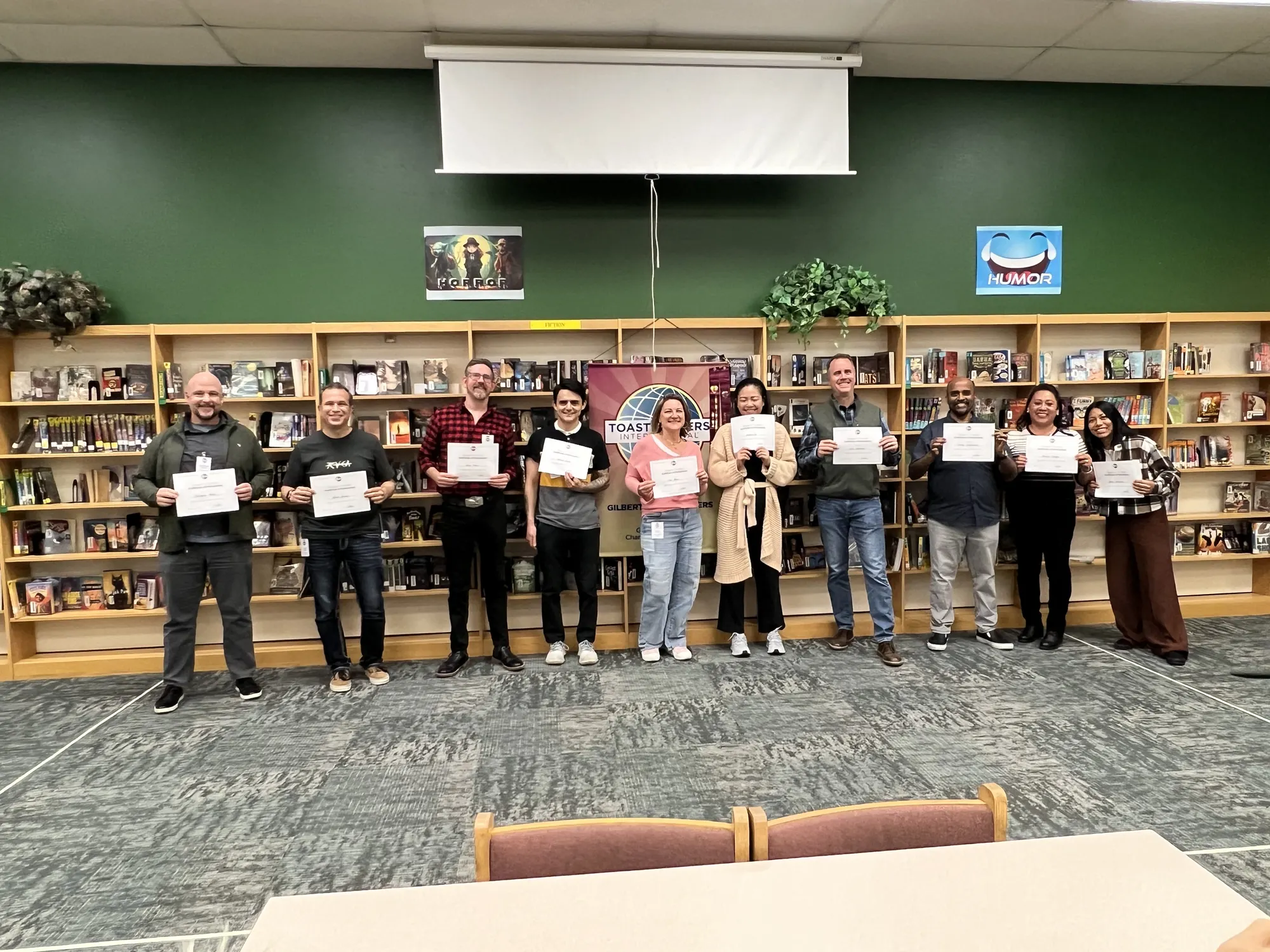 The contestants of the 2026 Gilbert Toastmasters Tall Tales contest lined up in front of the bookshelves, each holding their certificate